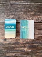 Two oracle cards with an ocean-themed design, displayed on a dark wood background. One card shows a blue, green, and beige beach gradient with the text 'May the Sea be your guide', and the other features a matching vertical beach design with the title 'Sea Oracle Cards' in gold.