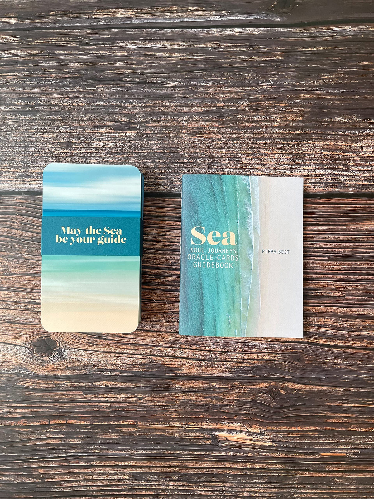 Two oracle cards with an ocean-themed design, displayed on a dark wood background. One card shows a blue, green, and beige beach gradient with the text 'May the Sea be your guide', and the other features a matching vertical beach design with the title 'Sea Oracle Cards' in gold.