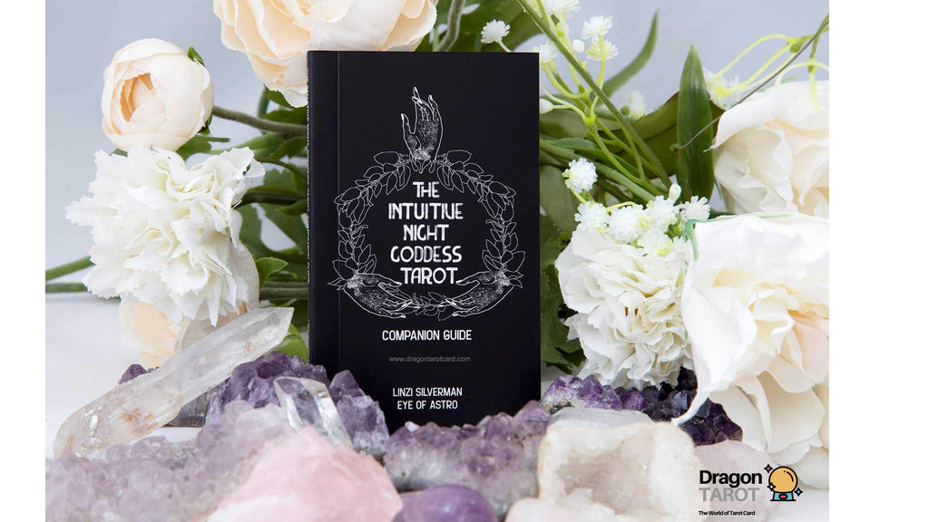 The Intuitive Not Godess Tarot Companion Guide book by Linda Severman, displayed upright amidst white roses, purple crystals, and clear quartz crystals.