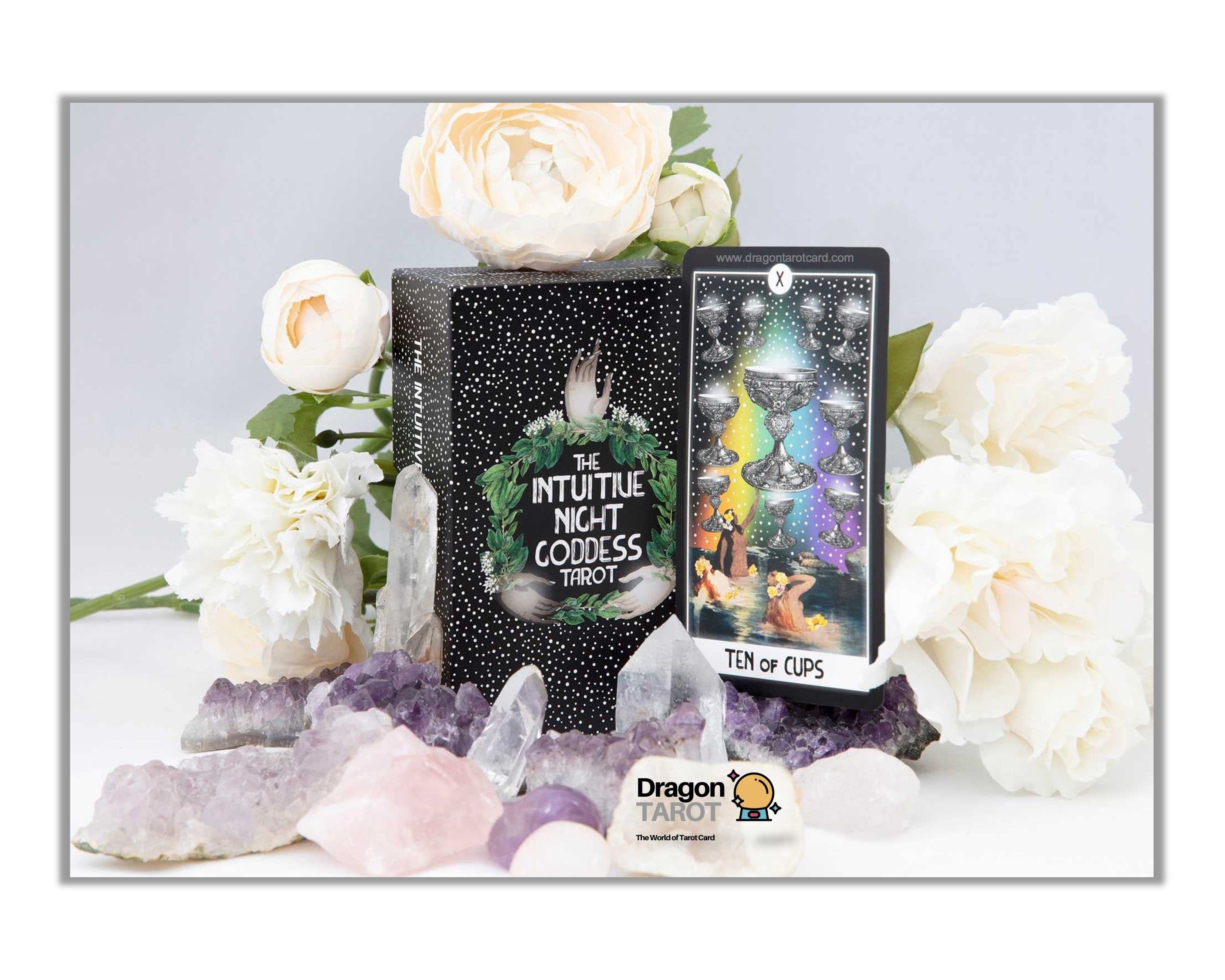 A boxed card deck and a single card displayed on a white surface with decorative white flowers, purple crystals, and a Dragon's Eye logo at the bottom. The box reads 'Nyttilte Night Goddess' and the card shows a colorful illustrated design.