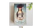 The 'Oracle of Mystical Moments' card deck box by Carina Wade-Stein, displayed on a light gray background with a decorative green leaf sprig in the top right corner. The box features an illustration of a woman in a dark hat and lace dress against a light blue background.