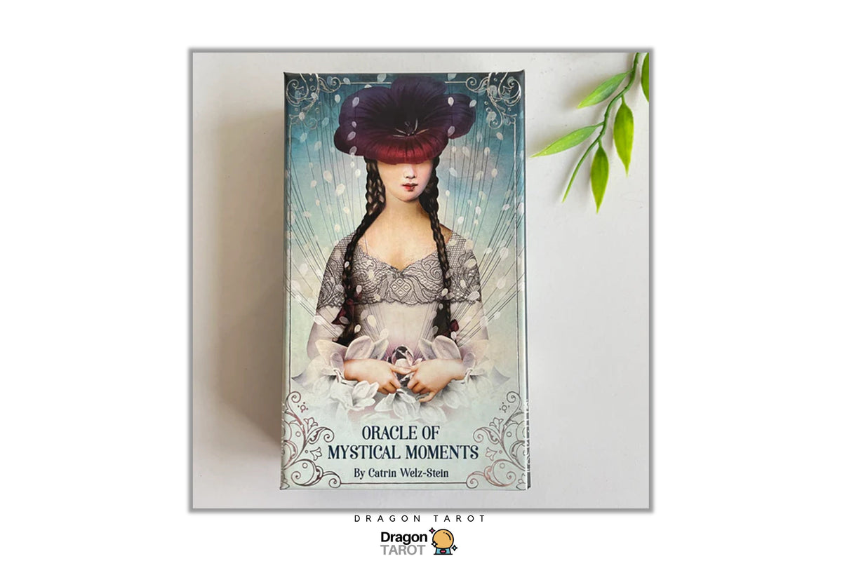 The 'Oracle of Mystical Moments' card deck box by Carina Wade-Stein, displayed on a light gray background with a decorative green leaf sprig in the top right corner. The box features an illustration of a woman in a dark hat and lace dress against a light blue background.