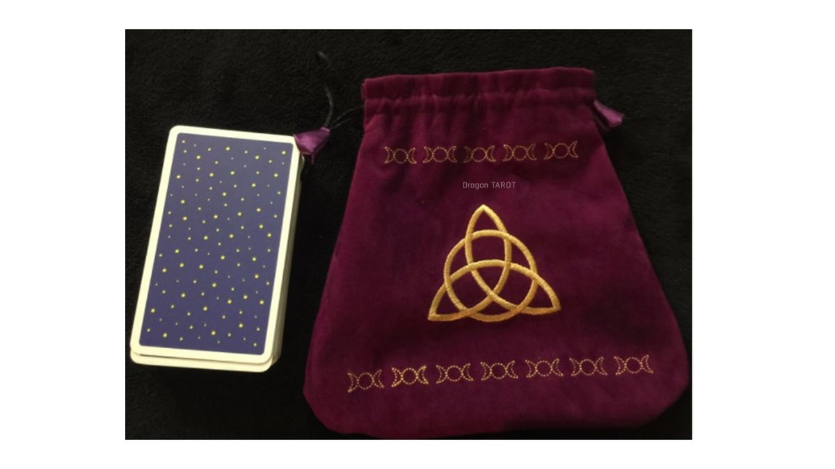 A tarot deck with a blue-backed card and a deep burgundy velvet drawstring pouch featuring gold embroidery of a triquetra knot and the word 'DEORATO' on a black background.
