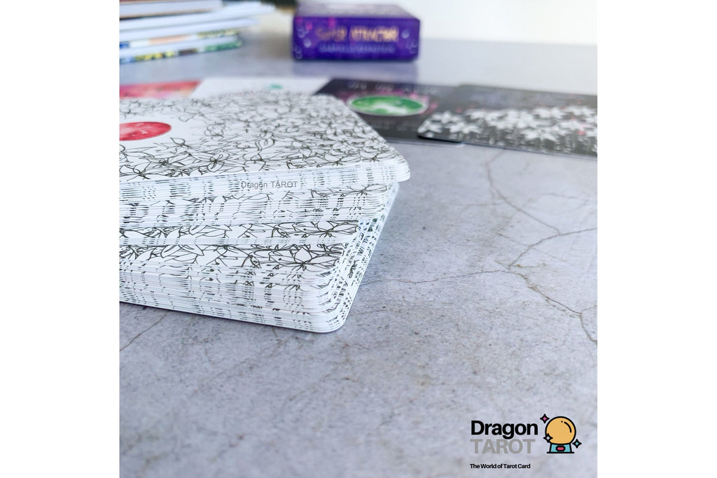 A stack of Dragon Tarot cards with a black and white floral patterned cover sits on a gray stone surface, with other cards and a purple box blurred in the background.
