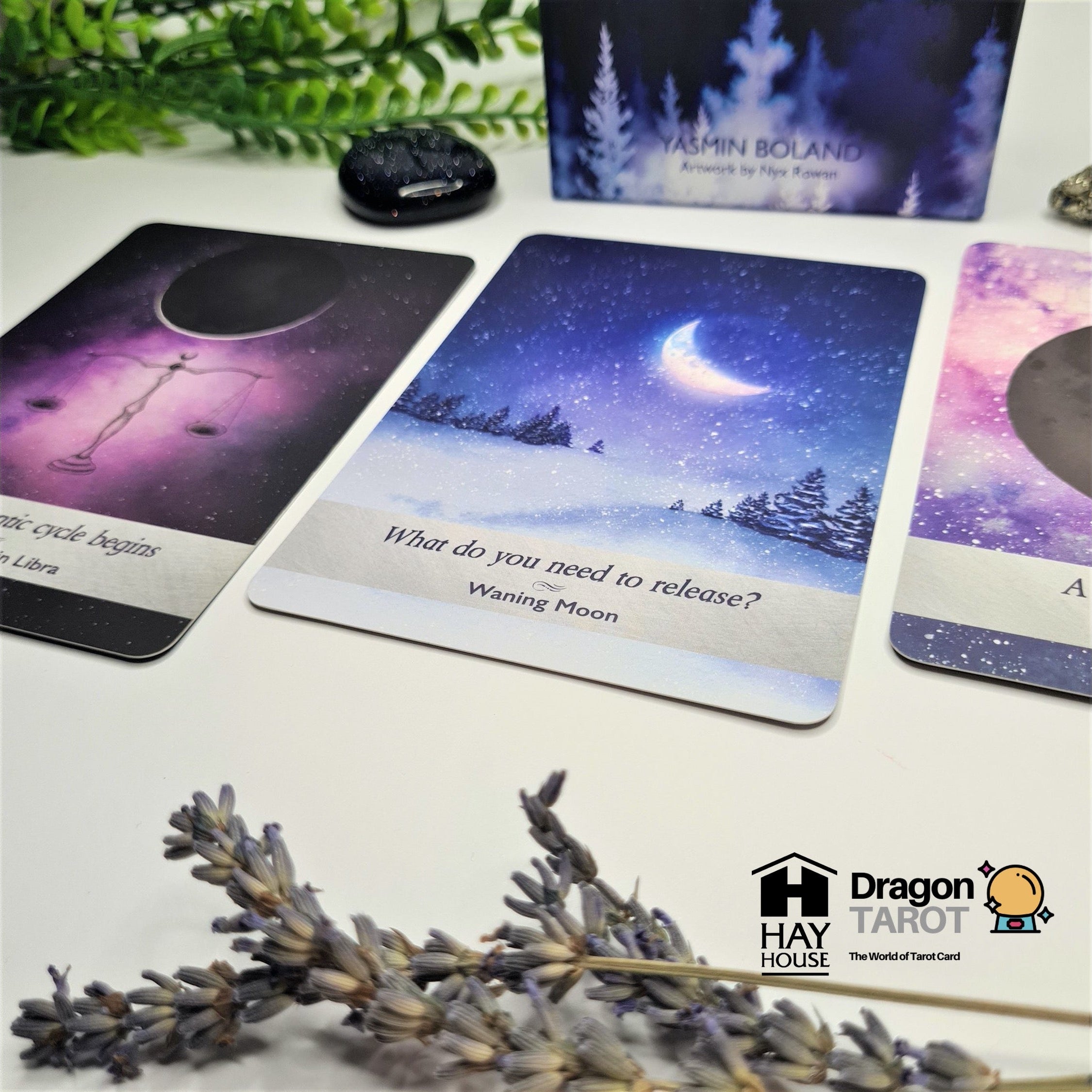 A tarot card deck set displayed on a light surface with decorative dried lavender and greenery. The main visible product is the tarot card deck, including cards and a box, with one central card showing a night sky and moon illustration.