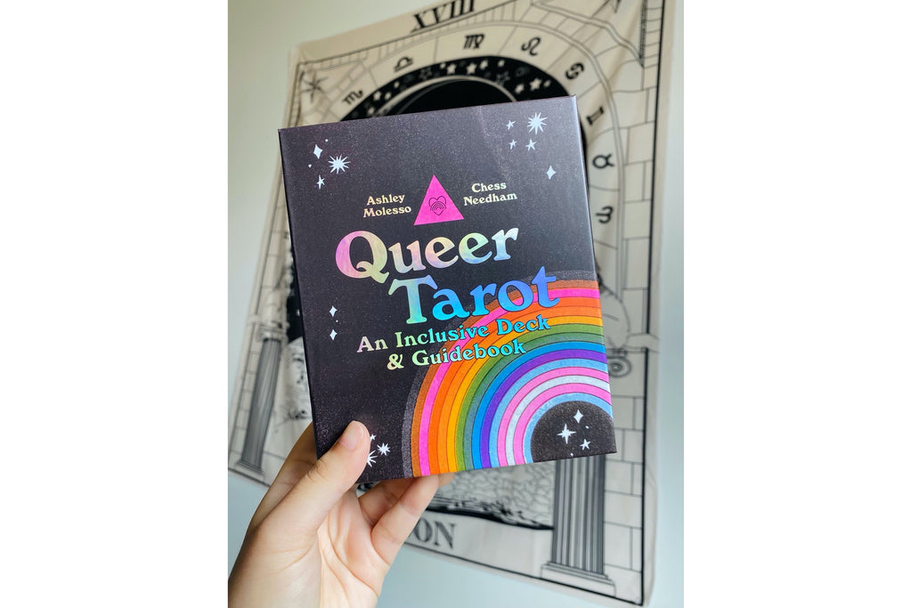 A hand holds a book titled "Queer Tarot: An Inclusive Deck & Guidebook" against a light gray background with a sketch of an archway and clock face.