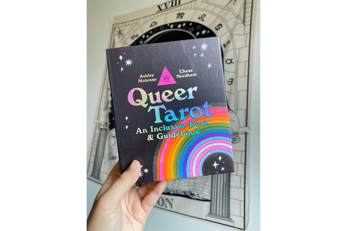 A hand holds a book titled "Queer Tarot: An Inclusive Deck & Guidebook" against a light gray background with a sketch of an archway and clock face.