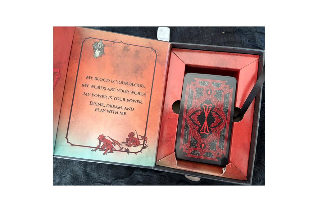 A boxed deck of playing cards shown open, with the red card back and a red inner tray visible, alongside an open illustrated insert page with text and artwork. The product is photographed on a dark fabric surface.