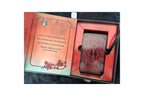 A boxed deck of playing cards shown open, with the red card back and a red inner tray visible, alongside an open illustrated insert page with text and artwork. The product is photographed on a dark fabric surface.