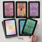 A set of tarot cards displayed on a light gray surface, including several face-up cards showing illustrated designs and a stack of cards with an iridescent back. A hand is holding one of the face-up cards.