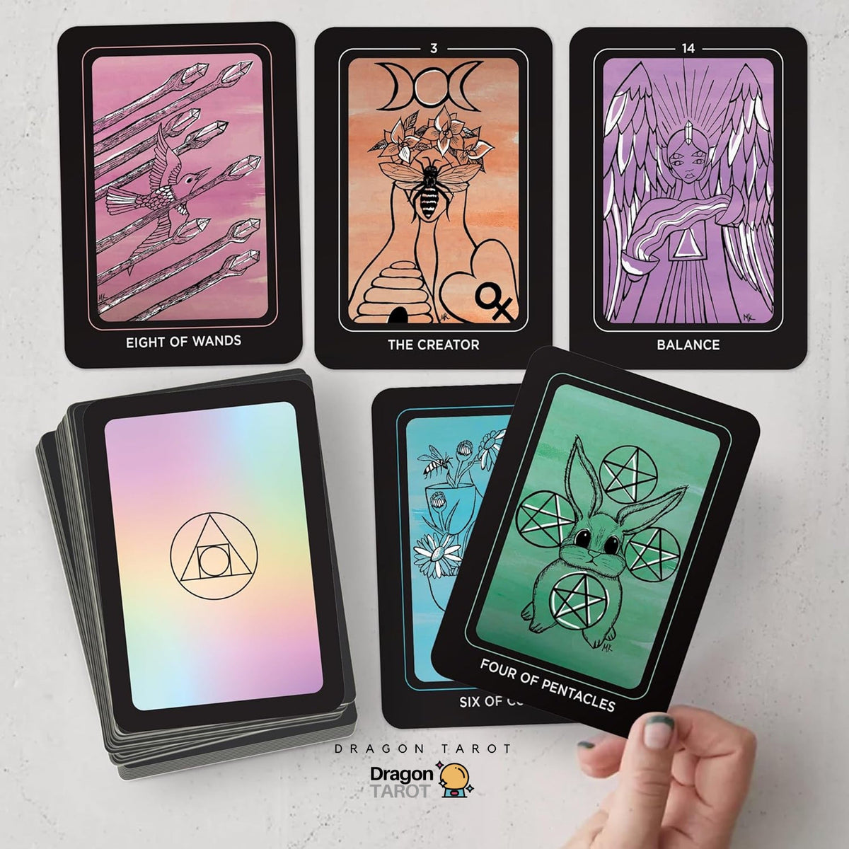 A set of tarot cards displayed on a light gray surface, including several face-up cards showing illustrated designs and a stack of cards with an iridescent back. A hand is holding one of the face-up cards.