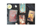 A tarot card deck with several illustrated cards laid out on a dark fabric surface, alongside small crystals and a round decorative object.