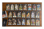 A tarot card deck displayed on a wooden board with multiple cards laid out in rows, a deck of cards standing upright on the left, and a black booklet or box at the bottom left. The cards feature colorful illustrated figures and scenes.