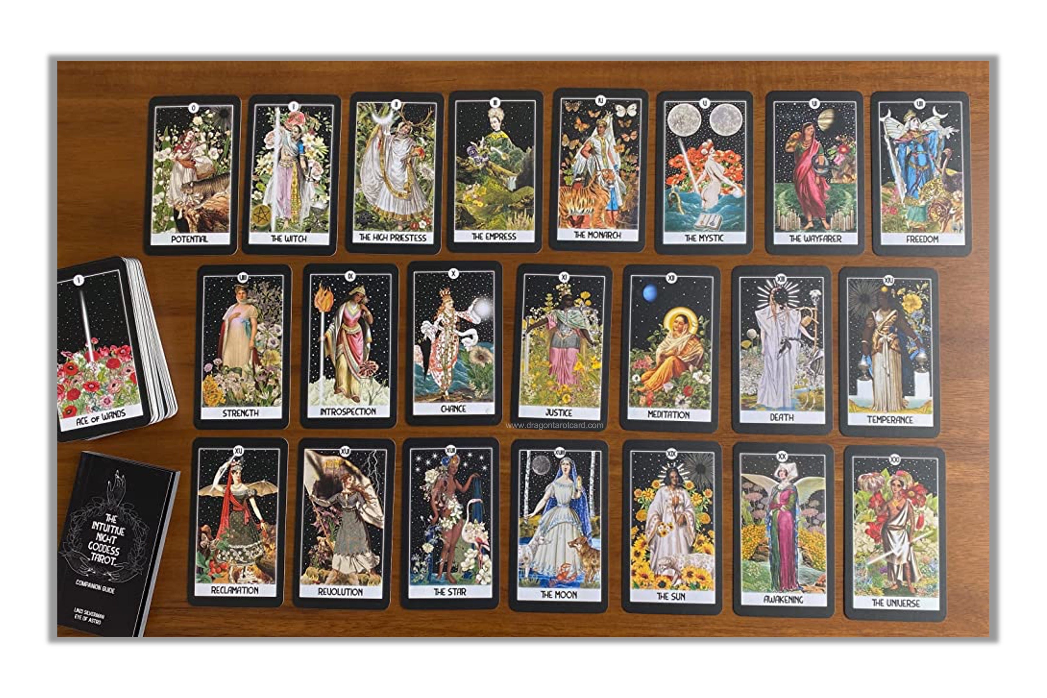 A tarot card deck displayed on a wooden board with multiple cards laid out in rows, a deck of cards standing upright on the left, and a black booklet or box at the bottom left. The cards feature colorful illustrated figures and scenes.