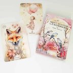 A Sweet Blossom Lenormand deck box and two illustrated cards displayed on a white background. The box features pink floral artwork with birds, and the cards show whimsical watercolor illustrations including a fox and a child.