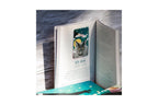 An open book with a small illustrated card or bookmark featuring a cat and the text '23 ASH', placed on a teal patterned surface against a dark wood background.