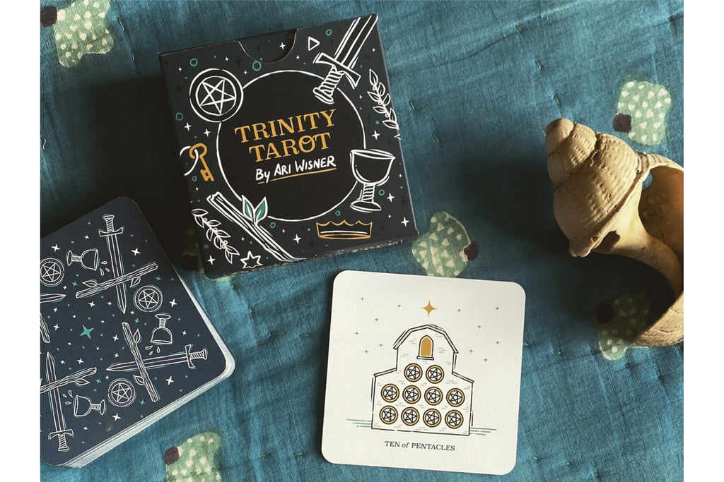 Trinity Tarot deck by Ari Werner, featuring a black box with white and gold celestial illustrations, a blue card tin, and a sample card on a textured blue fabric background with decorative seashell props.