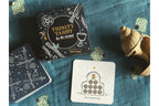 Trinity Tarot deck by Ari Werner, featuring a black box with white and gold celestial illustrations, a blue card tin, and a sample card on a textured blue fabric background with decorative seashell props.