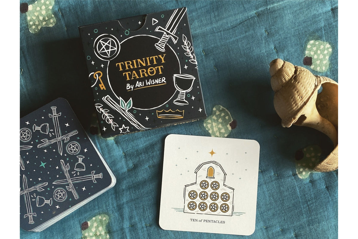 Trinity Tarot deck by Ari Werner, featuring a black box with white and gold celestial illustrations, a blue card tin, and a sample card on a textured blue fabric background with decorative seashell props.