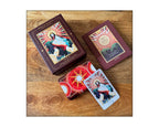 A boxed card deck and two cards displayed on a wooden surface. The packaging and card backs feature a dark reddish-brown border with a central religious illustration of a haloed figure and a red sunburst design.
