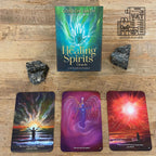 Healing Spirits Oracle card deck box by Gordon Smith displayed on a wooden surface with two dark crystals and three illustrated oracle cards below. The box features a green-to-blue gradient with glowing hands artwork, and the cards show colorful spiritual imagery.