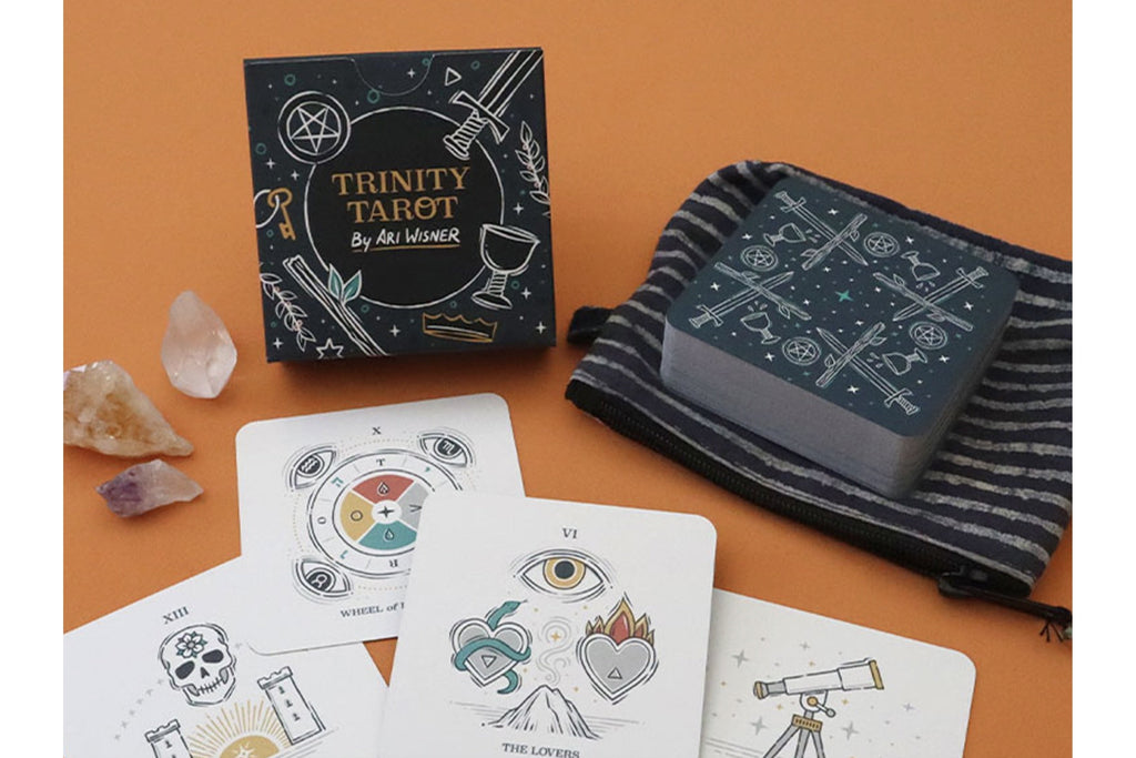 Trinity Tarot deck by Ali Memon displayed on an orange background, including the black illustrated box, a striped drawstring bag, and several white cards with line-art illustrations.