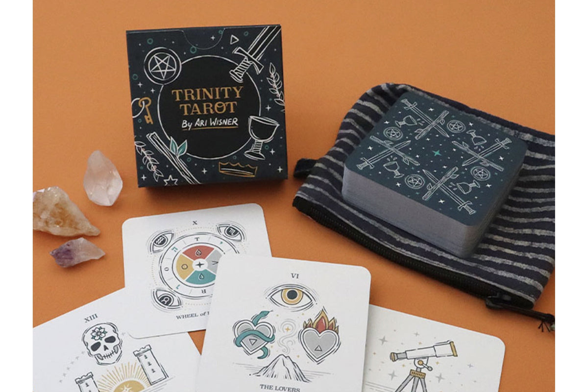 Trinity Tarot deck by Ali Memon displayed on an orange background, including the black illustrated box, a striped drawstring bag, and several white cards with line-art illustrations.