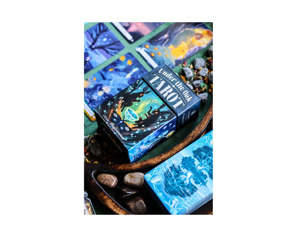 A boxed tarot deck with a dark blue illustrated box labeled 'The Fool Tarot' sits on a rustic tray beside matching tarot cards and small stones.