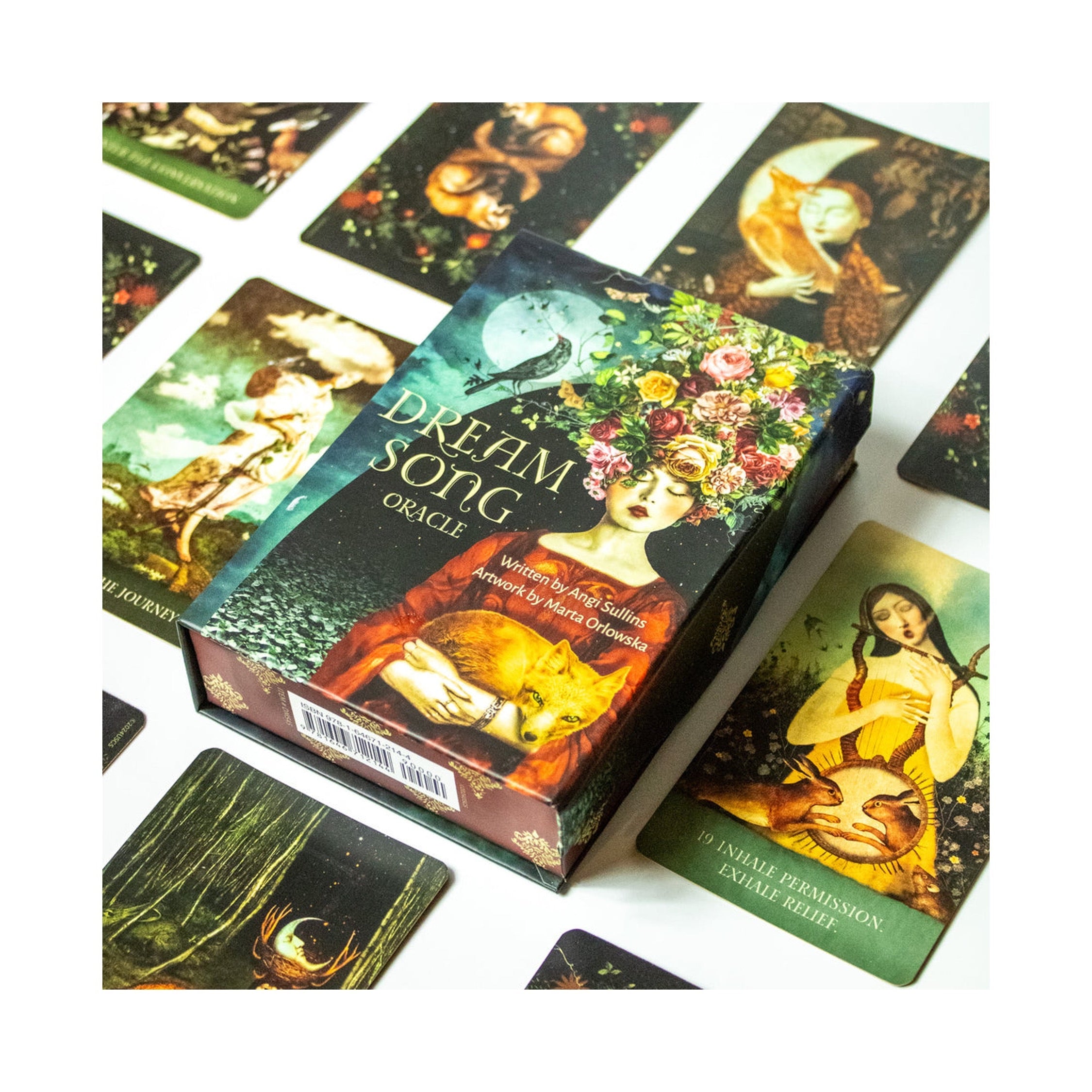 A boxed card deck titled "Dreaming Song" is shown on a white surface with several illustrated cards arranged around it. The box features fantasy-style artwork with a woman and warm glowing tones, and the cards also have detailed printed illustrations.