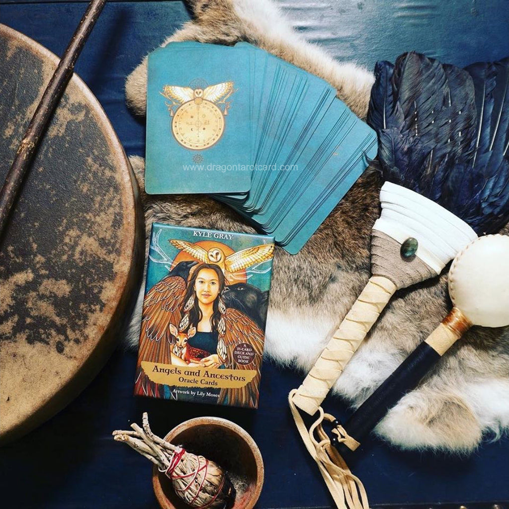 A styled flat-lay of a tarot deck, featuring a fanned deck of teal cards, a single upright card depicting a woman with a horned headpiece, and various mystical props including a drum, a selenagins ritual tool, a small bowl with a sprig, and a dark blue fabric backdrop.