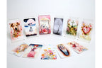 A set of illustrated oracle or tarot-style cards displayed on a white background, with several cards standing upright in clear acrylic holders and others fanned out in front. The cards feature pastel watercolor-style artwork including a dog, a woman under a pink umbrella, floral scenes, a tree, a cross, and a heart motif.