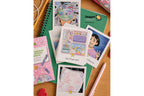 A styled flat-lay of Dragon Tiger themed stationery items on a wooden desk background, including a green Dragon Tiger notebook, a pink 'Everyday Organizer' book, and several illustrated polaroid-style cards.