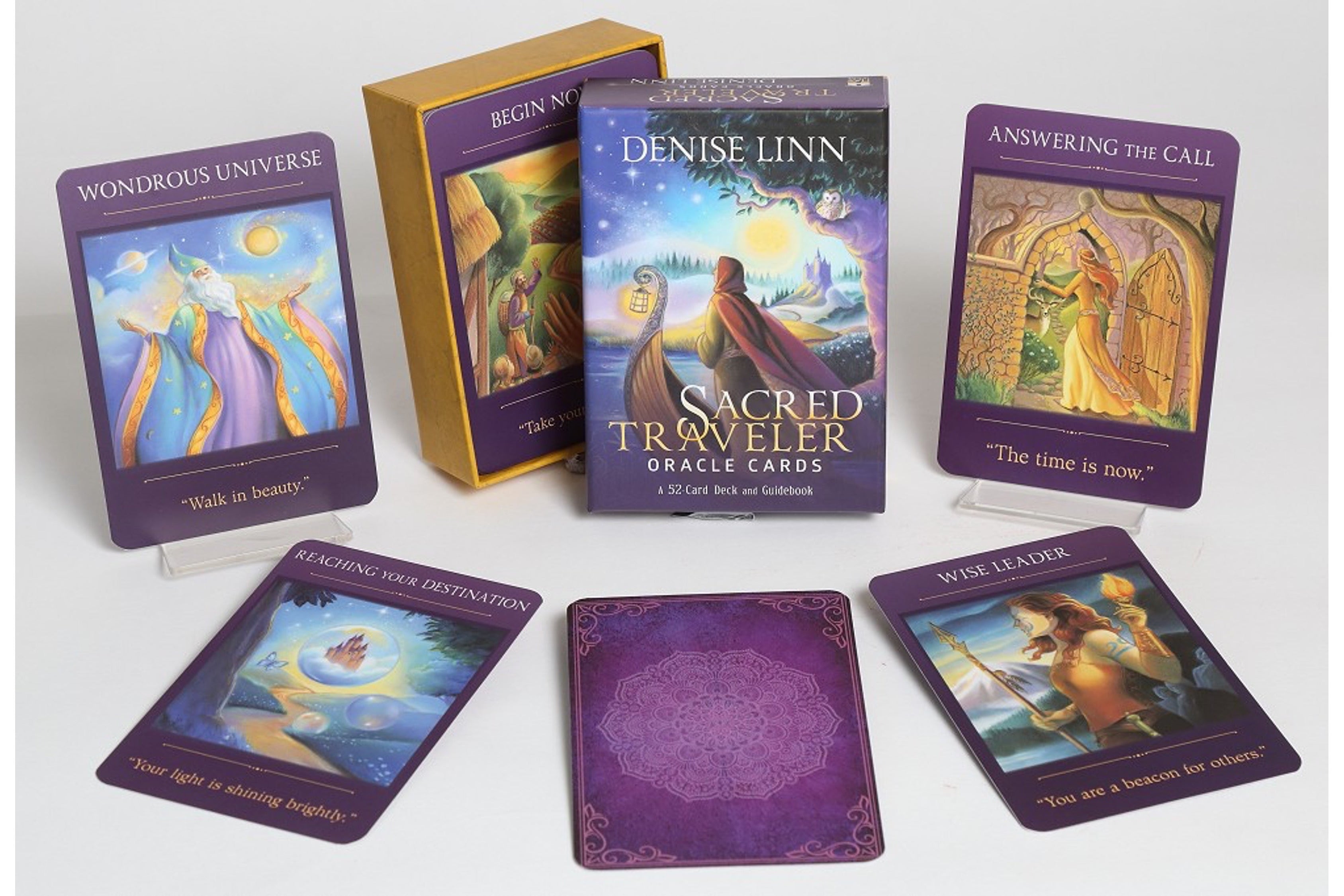 Sacred Traveler Oracle Cards deck by Denise Linn, showing the main box, two purple-bordered card fronts, two card backs, and a gold booklet on a light gray background.